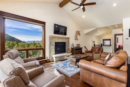 A living room with a large window offering a mountain view. Features include a stone fireplace, mounted TV, brown leather and fabric sofas, a wooden coffee table, and a ceiling fan.