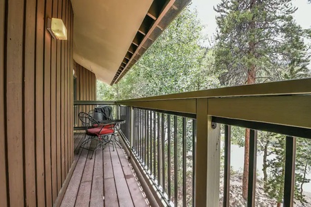 A narrow wooden balcony with a black metal railing features two metal chairs with red cushions and a small table. Trees are visible in the background.