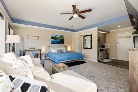 A spacious, well-lit bedroom with a ceiling fan, a bed with blue bedding, a sofa, and a small kitchenette area. There is a wall-mounted television and an open entrance to a bathroom or closet area.