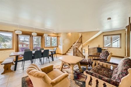 Spacious living and dining area featuring a wooden table with black chairs, cozy armchairs, a coffee table, and large windows offering scenic views. A staircase is visible in the background.