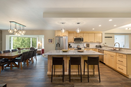 A modern kitchen-dining area with light wood cabinets, stainless steel appliances, a central island with three bar stools, and a dining table with eight chairs beneath contemporary light fixtures.