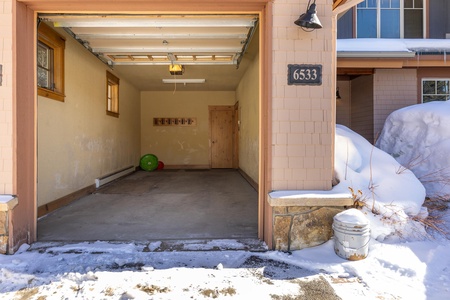 An open garage of a residential home showing an empty interior with a few items on the concrete floor and snow outside. The house number "6533" is visible above the garage entrance.