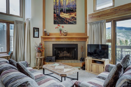 A living room with a large fireplace, TV, and a sofa with patterned upholstery. Windows on either side of the fireplace reveal an outdoor view. A coffee table is centered in front of the sofa.