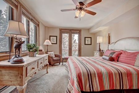 A bedroom with a striped bedspread, wooden furniture, two table lamps, a ceiling fan, and large windows with a view of trees outside.