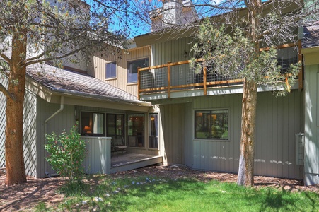 Two-story gray house with a wooden balcony and a small porch. The house is surrounded by trees and has a grassy backyard.