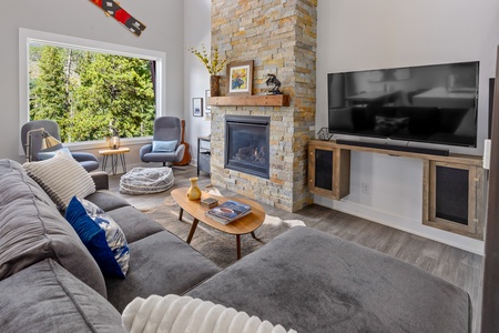 A modern living room with a gray sectional sofa, stone fireplace, wall-mounted TV, and large window overlooking trees. The room features wooden accents and a decorative canoe on the wall.