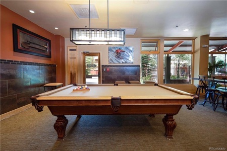 A room with a pool table in the center, art on the walls, a large pendant light above the table, and windows in the background showing trees and outdoor seating.