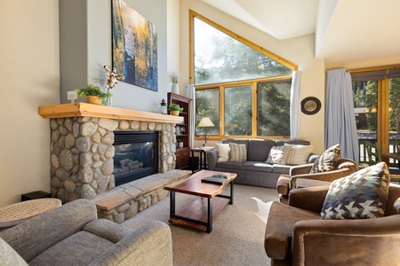 A cozy living room features large windows, a stone fireplace, two gray sofas, two leather armchairs, a wooden coffee table, and a bookshelf. Sunlight filters through the trees outside.