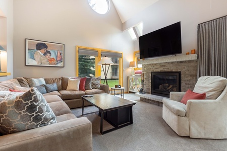 Living room with beige sectional sofa, patterned pillows, coffee table, armchair, brick fireplace, wall-mounted TV, large windows, and artwork on wall. Natural light illuminates the space.