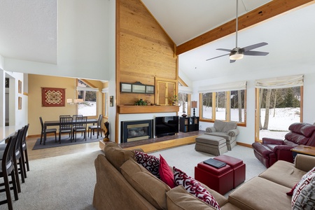 Spacious living room featuring a sectional sofa, armchair, fireplace with wooden mantel, and large windows with views of a snowy landscape. Adjacent dining area with a table and chairs is visible.