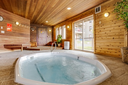 An indoor hot tub with bubbling water is set in a wood-paneled room with large windows, a bench, coat hooks, and a plant in the corner.