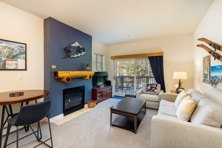 A cozy living room with a sofa, coffee table, TV, fireplace, and a small dining area. The room has a blue accent wall and sliding glass doors leading to a balcony with a view of greenery.