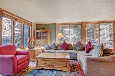 A living room with large windows, a gray L-shaped sofa adorned with cushions, a red armchair, wooden furniture, and a colorful rug.