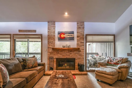 A cozy living room features a stone fireplace with a Colorado-themed painting above it, a brown sofa, an armchair, and a large window with a view of a wooded area.