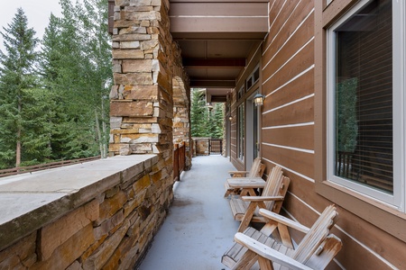 A wooden balcony with stone pillars features three wooden chairs and overlooks a forested area. The balcony has wooden siding and a wall lamp.