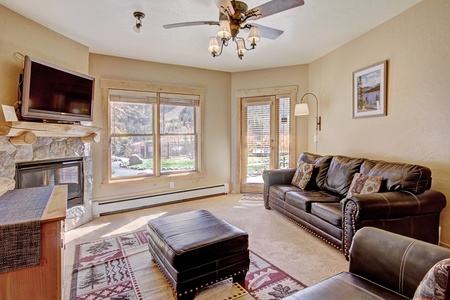 A cozy living room with a leather sofa, ottoman, stone fireplace, wall-mounted TV, and large windows offering a scenic view. A ceiling fan and floor lamp provide additional light. .