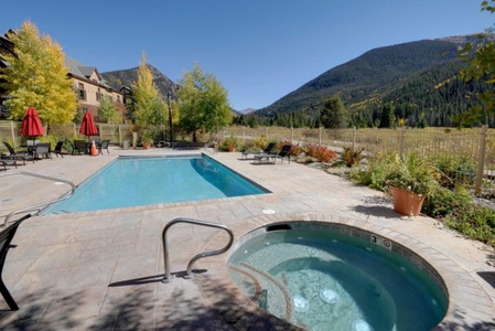 Outdoor pool and hot tub on a sunny day with mountain and forest views in the background. Red umbrellas and lounge chairs are set around the pool area.