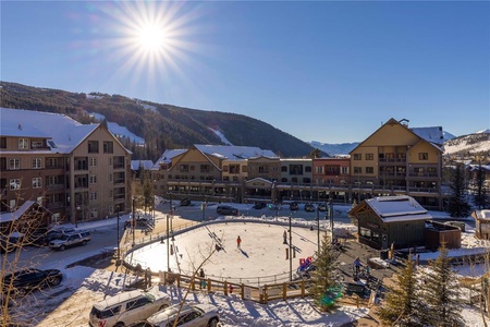 A sunny day with people ice skating in an outdoor rink surrounded by buildings and snow-covered mountains in the background.