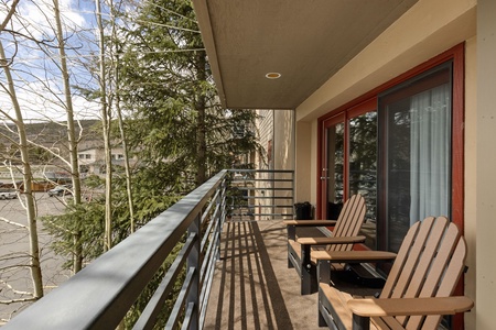 Balcony with two Adirondack chairs overlooking a parking area and trees, with a view of neighboring buildings in the background.