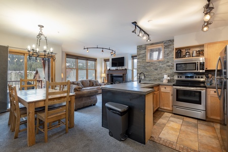 A modern open-plan kitchen and living area with wooden cabinetry, stainless steel appliances, a stone accent wall, a dining table with chairs, and a cozy seating area with a brown sofa and large windows.