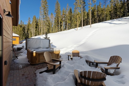 A snow-covered outdoor patio with wooden chairs and a hot tub near a cabin in a forested area under a clear blue sky.