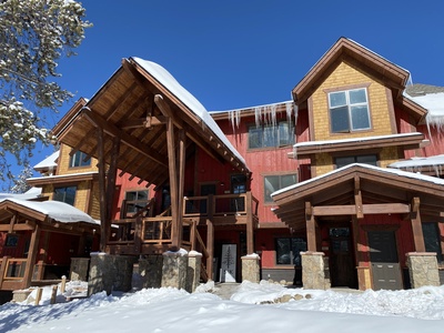 A red and yellow wooden house with steep roofs, covered in snow and icicles, set against a clear blue sky.