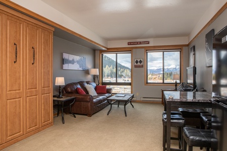 A cozy living room with a brown leather couch, two windows offering mountain views, and a wall-mounted TV. The room has wooden accents, a small dining area, and a wall-mounted cabinet.