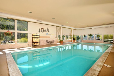 An indoor swimming pool area with large windows offering an outdoor view, potted plants, a wall mural, and a seating area with a table and chairs.