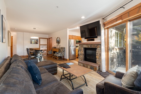 A living room with a sectional sofa, coffee table, and fireplace with a mounted TV. An open kitchen and dining area are in the background, and sliding glass doors open to an outdoor patio.