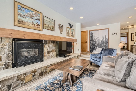 Cozy living room with stone fireplace, wooden coffee table, blue armchair, and beige sofa. Walls adorned with paintings and antler decor. Rug on floor and door in the background.