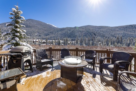 A wooden deck with patio furniture, a fire pit, and a barbecue grill offers a scenic view of snow-covered mountains and pine trees under a clear blue sky.