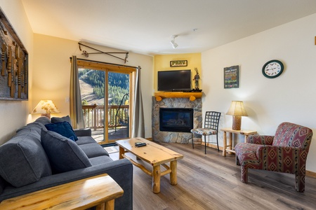 A cozy living room features a sofa, patterned armchair, wooden coffee table, fireplace, and TV. Sliding glass doors open to a balcony with a scenic view of a mountainous landscape.