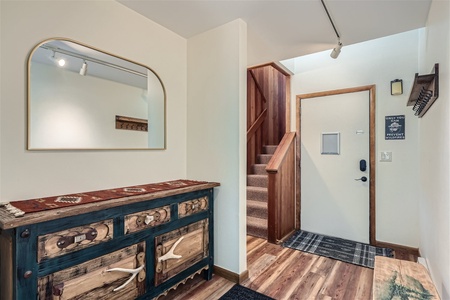 A hallway with a wooden sideboard featuring decorative items, a large mirror, a carpeted staircase, and a door with a small window. The floor is a mix of wooden planks and rugs.