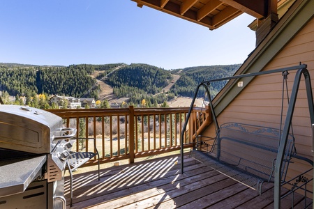 A wooden balcony with a grill and a metal bench swing overlooks a scenic forested mountain landscape on a clear, sunny day.