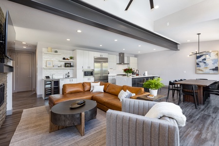 Modern open-concept living space featuring a kitchen with white cabinets, stainless steel appliances, a kitchen island, a tan sectional sofa, a gray armchair, a wooden dining table, and decor items.