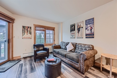 A cozy living room with a brown couch, a black armchair, an ottoman, and wooden furniture. Vintage ski-themed posters adorn the walls. Large windows provide views of an outdoor area.