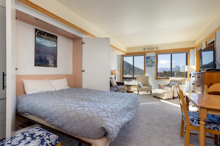 A well-lit studio apartment features a Murphy bed, chairs, a desk, and panoramic mountain views through large windows. A "Colorado" poster hangs above the bed.