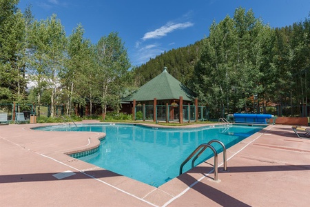 Outdoor swimming pool with clear blue water, surrounded by a concrete deck, lounge chairs, and trees. A green-roofed pavilion is visible in the background against a backdrop of wooded hills.