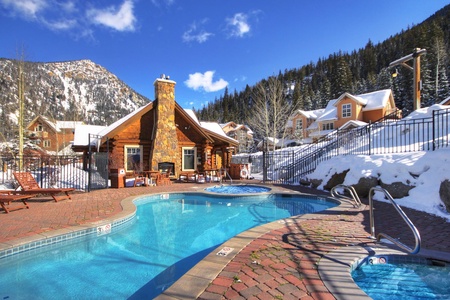 A lodge with a stone chimney near a snow-covered mountain features an outdoor swimming pool and hot tub, surrounded by a fence, with clear blue skies in the background.