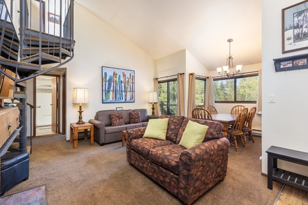 A living room with a patterned sofa and armchair, a dining area with a table and chairs, spiral staircase, wall artwork, lamps, and large windows showcasing greenery outside.