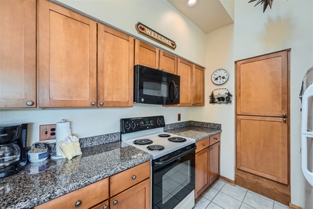 A kitchen with wooden cabinets, a granite countertop, a black stove, a black microwave, a coffee maker, a paper towel holder, a wall clock, and a "Welcome" sign above the cabinets.