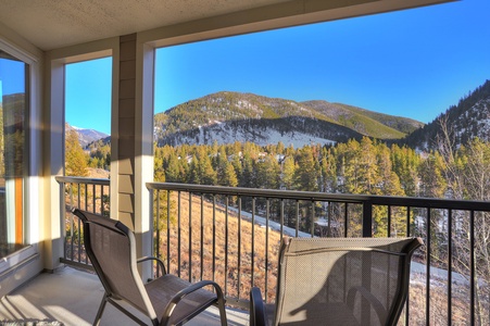 A balcony with two chairs overlooks a scenic mountain view featuring slopes covered with evergreen trees and patches of snow under a clear blue sky.