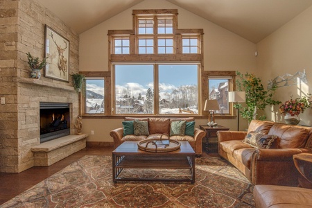 A cozy living room with a stone fireplace, leather sofas, a wooden coffee table, and a large window showcasing a snowy outdoor scene. The room is decorated with various plants and a patterned rug.