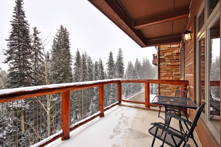 A snowy balcony with wooden railings and metal chairs overlooks a forest of tall pine trees.