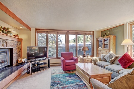 A cozy living room with a fireplace, TV, armchair, sofa, and a wooden coffee table. Large windows offer an outside view, and a colorful rug is placed on the carpeted floor.