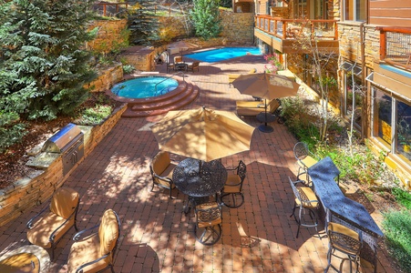 Outdoor patio with brick flooring, featuring a round dining table with umbrellas, cushioned chairs, a BBQ grill, a hot tub, and a swimming pool. Surrounding area includes trees and a stone wall.