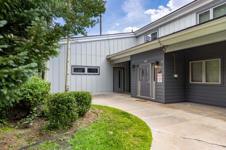 A modern building exterior with gray siding, large windows, and a covered entryway surrounded by greenery and a concrete walkway.