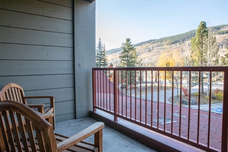 A small balcony with two wooden chairs overlooks a parking lot, trees, and distant mountains under a clear sky.