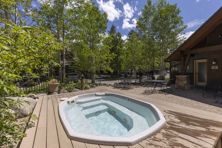 An outdoor hot tub surrounded by a wooden deck and greenery, with patio furniture and a fence visible in the background under a sunny sky.