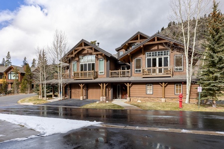 A modern, two-story wooden house with large windows, balconies, and a garage, set in a mountainous area on a cloudy day. A red fire hydrant is visible nearby. Snow is on the ground and partially melted.